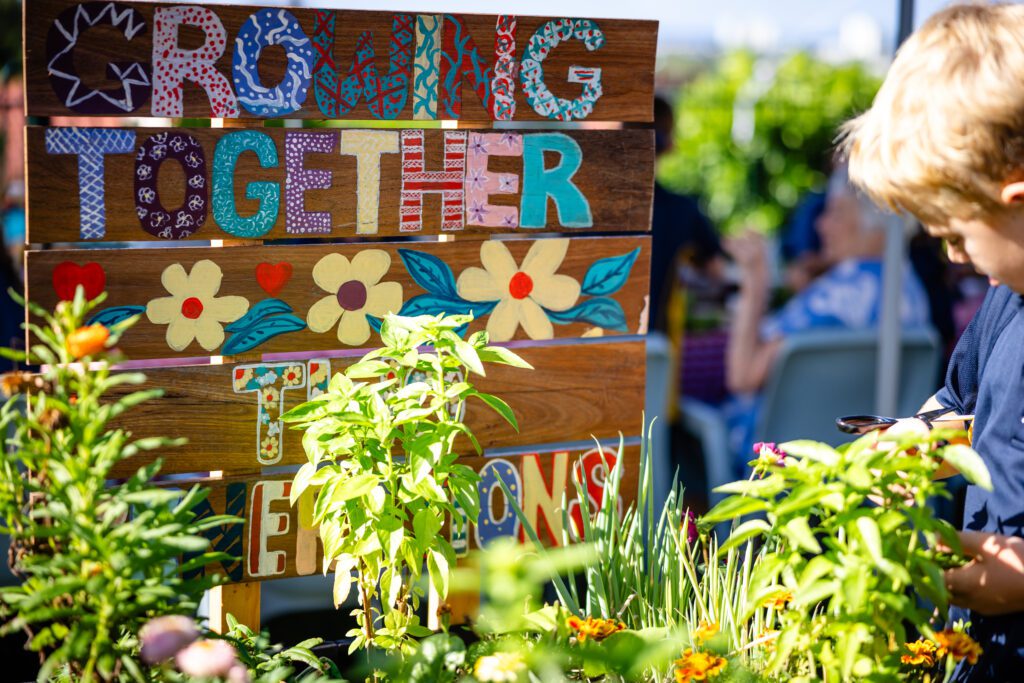 Hand creafted sign with multi-coloured letters amongst green plants.